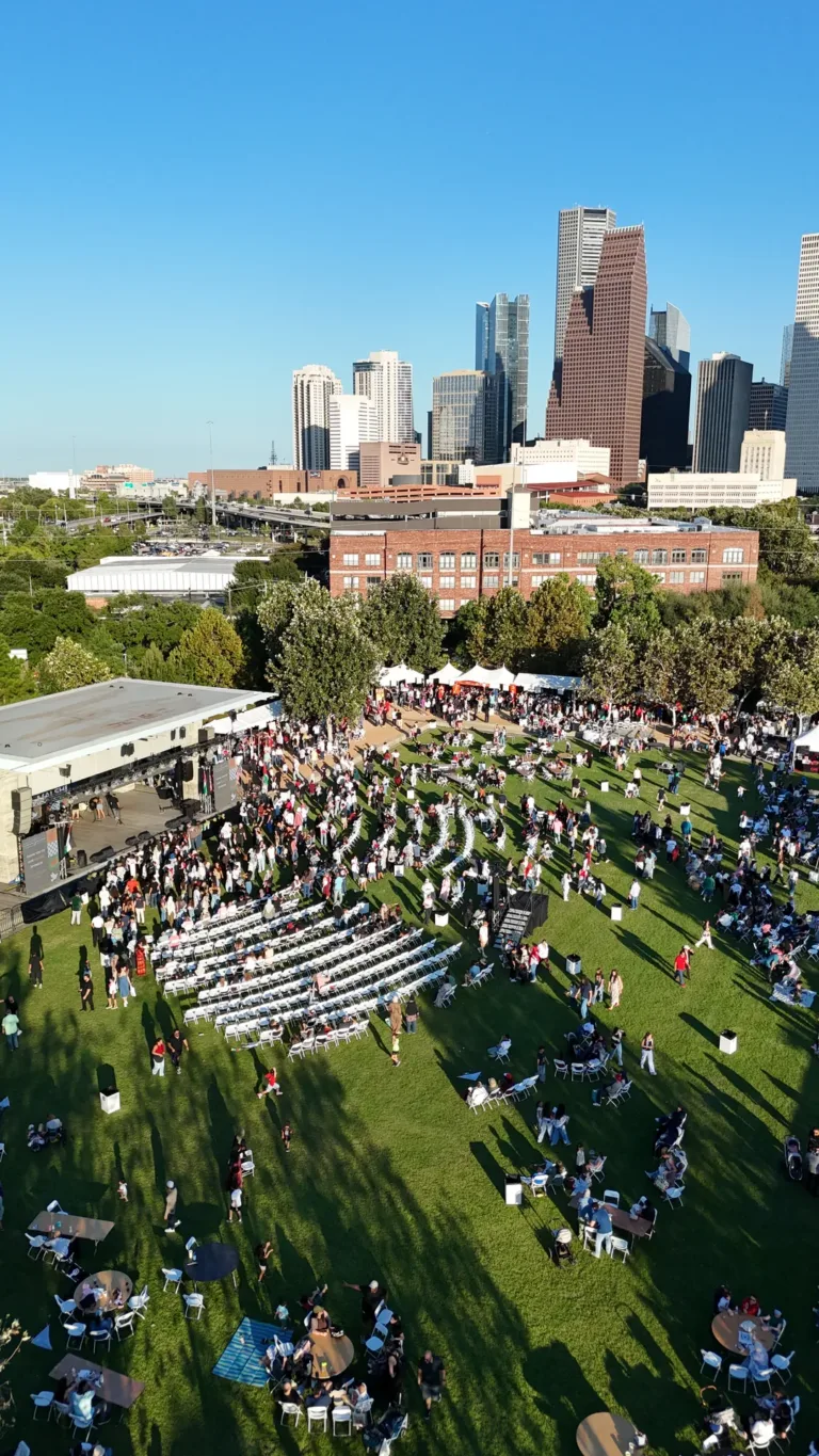 An aerial view shows people among rows of white chairs on a green lawn with tall city buildings beyond the trees.