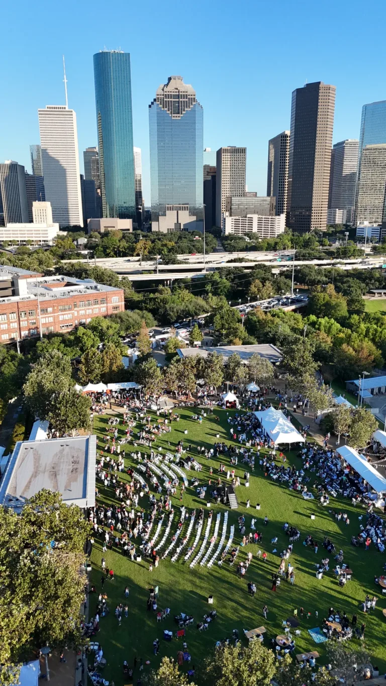 Rows of white chairs, people, and market tents fill a lawn beneath a skyline of tall buildings.