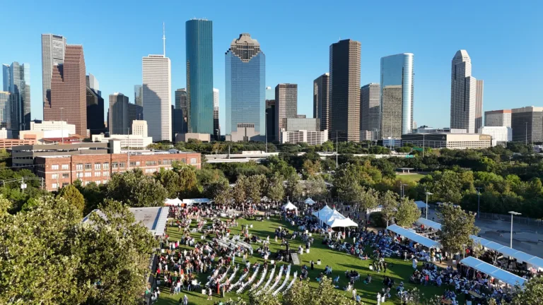 A wide aerial view frames city towers above a tree lined lawn with people, white chairs, and tents.