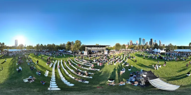 A curved panoramic view spans rows of white chairs, people on the lawn, a covered stage, and surrounding tents beneath a blue sky.