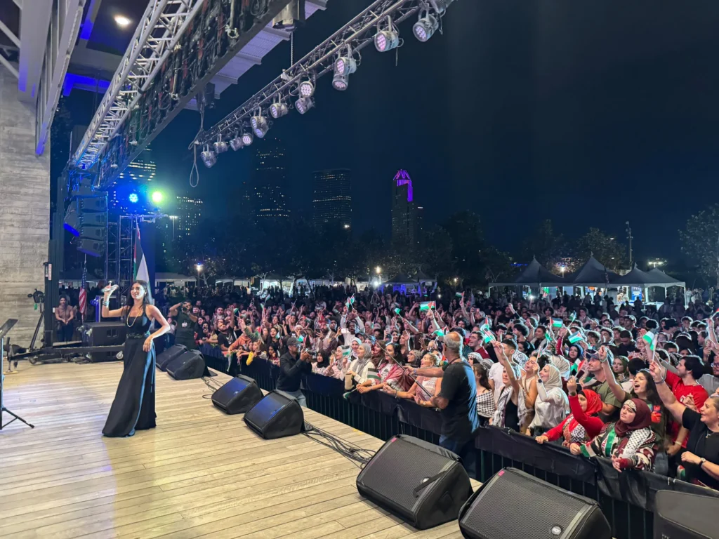 A singer performing beside a crowd waving Palestinian flags, with the Houston skyline at night.