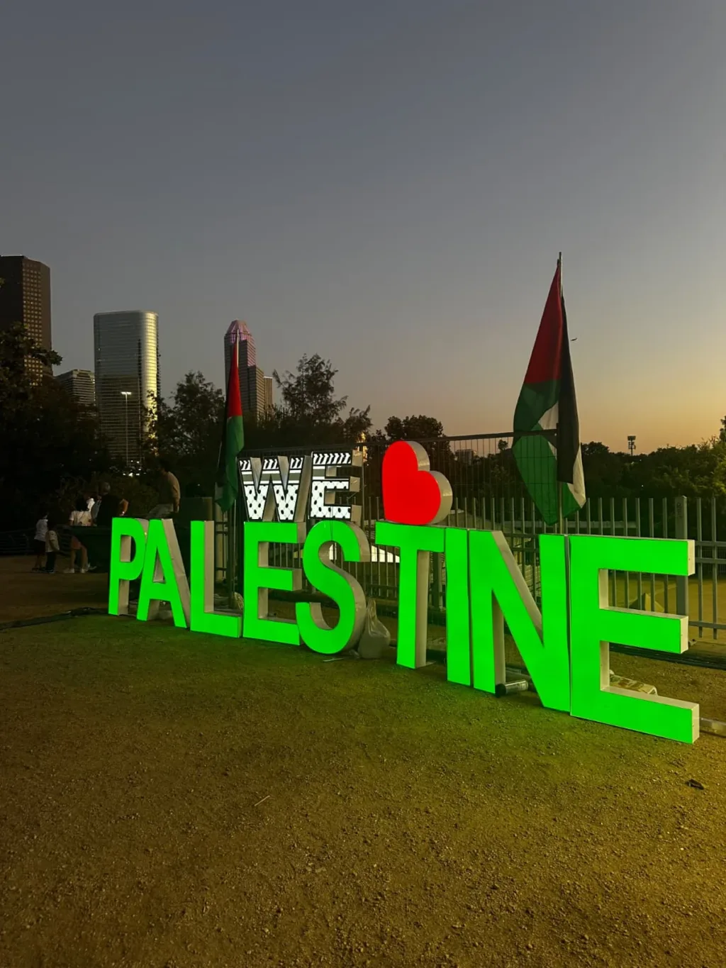 Illuminated Palestine lettering and Palestinian flags at dusk, with the Houston skyline behind the festival grounds.