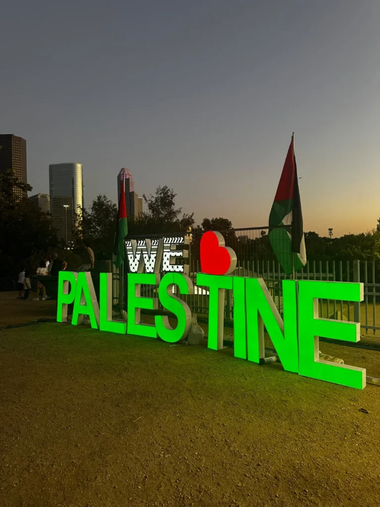 Illuminated Palestine lettering and Palestinian flags at dusk, with the Houston skyline behind the festival grounds.